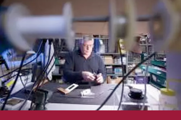 Man standing behind a desk surrounded by electrical wires and coils; he is concentrating on repairing a tool