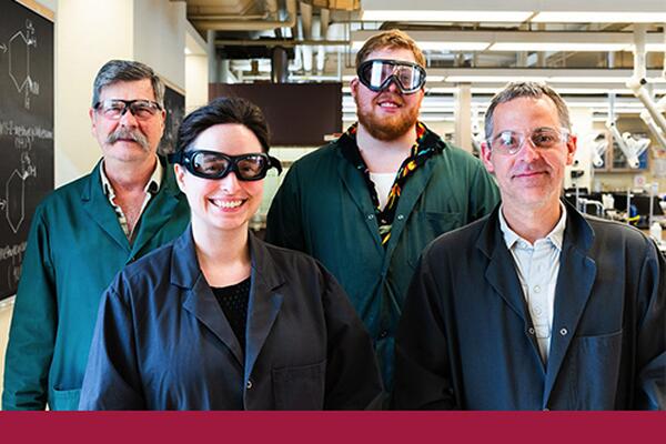 Group of chemistry staff and faculty members wearing safety goggles smile into the camera