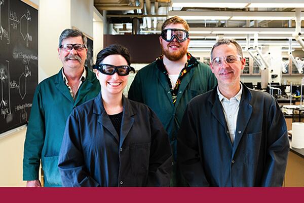 Group of faculty and staff wearing lab coats and goggles face the camera and smile