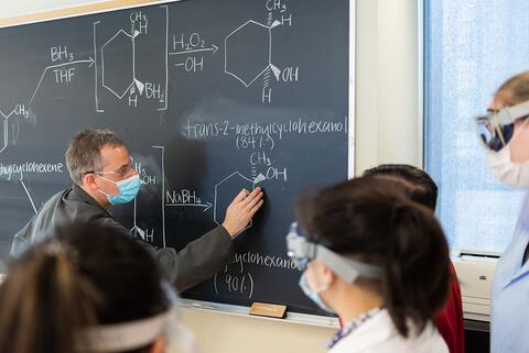 Students watch a faculty member write a Chemistry formula on a chalkboard