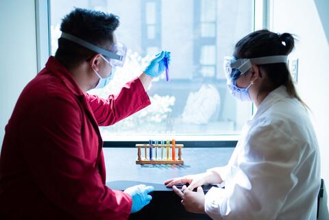 Two students wearing protective equipment look at a vial filled with liquid chemical