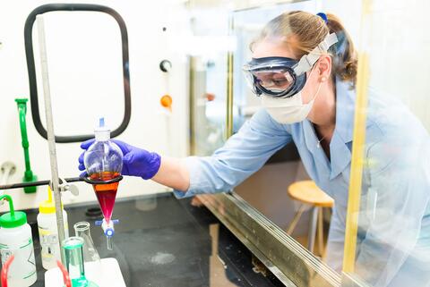 Student wearing protective equipment, including a face mask, goggles, and gloves, handles a flask with chemical liquid as part of an experiment