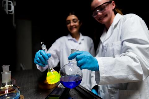 Two students wearing lab coats and goggles hold flasks of chemical liquid