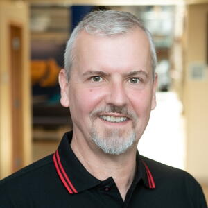 Head shot of Dr. Dominik Barz, man with grey hair, brown eyes, smiling in a blue collared shirt